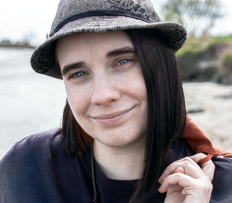 Bild der Autorin Kathrin Stoll, sie trägt ihren Hut als Markenzeichen, hat graublaue Augen. Die Haare sind zu einem Longbob geschnitten. Haarfarbe: Oberhaar: Dunkelbraun, Unterhaar: Kupferrot. Sie trägt ein schwarzes T-Shirt